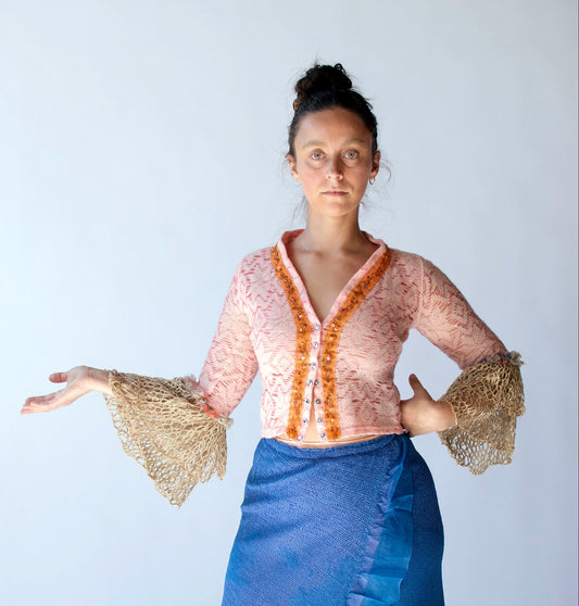A woman modeling a two-piece outfit consisting of a peach lace top with orange trim and textured beige bell sleeves, paired with a blue ruffled skirt, set against a plain light gray background.