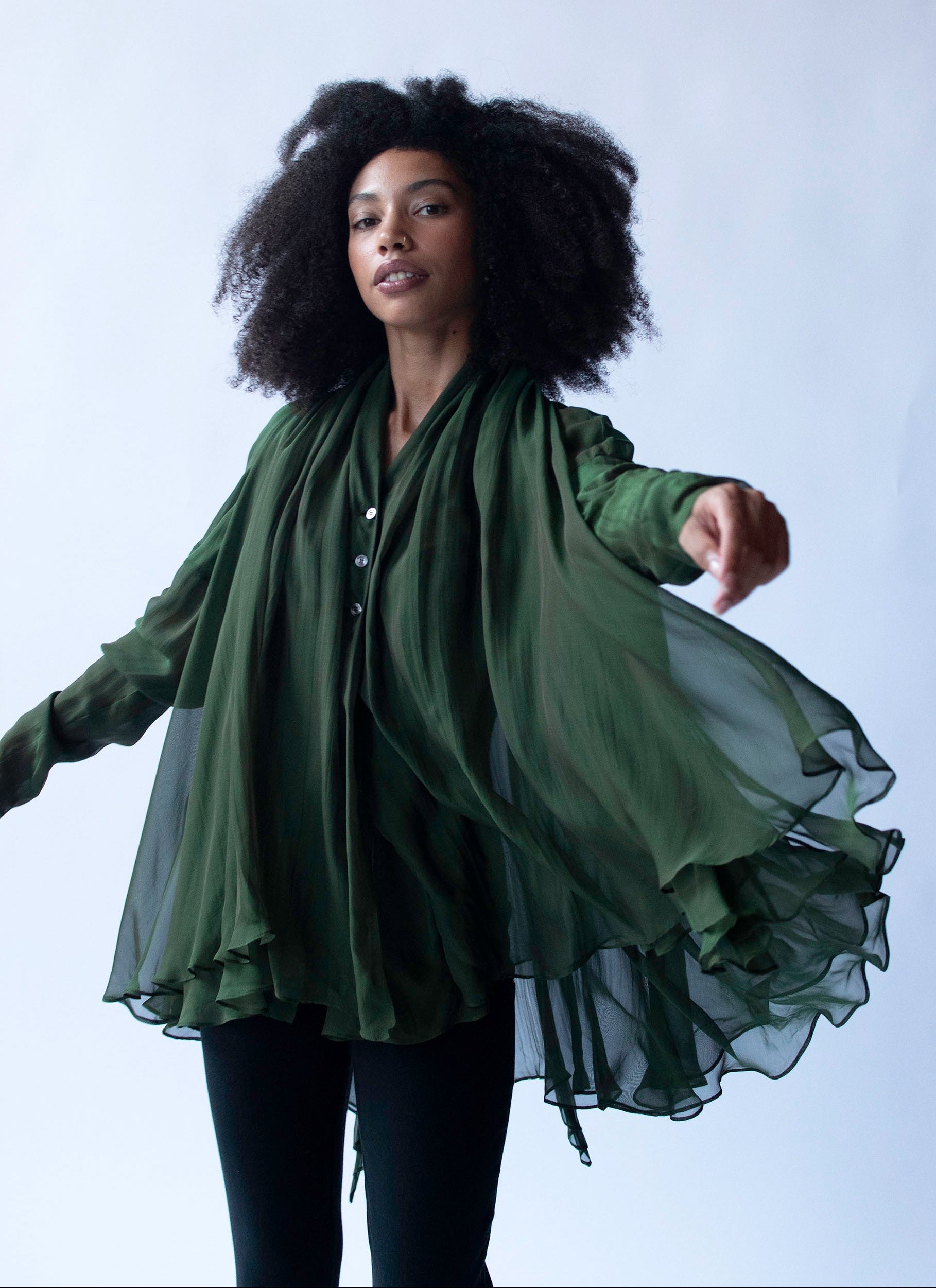 A woman with voluminous dark curly hair wearing a flowing forest green sheer long-sleeve shirt and matching draped scarf, posed against a light gray studio background.